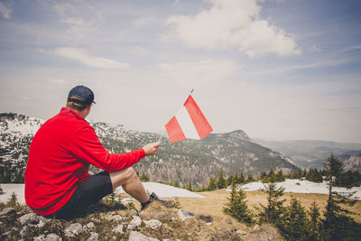 Rear view of man holding flag while sitting on mountain against sky