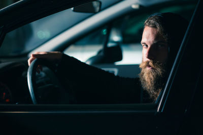 Portrait of bearded young man sitting in car seen through window at night