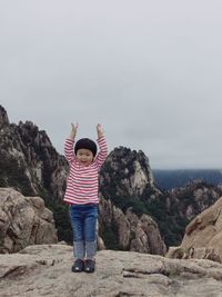 Full length of cute boy with arms raised standing on rock against sky