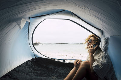 Side view of smiling mature woman using phone in tent at beach