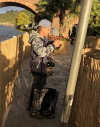 Side view of man photographing with camera while standing on footpath in city