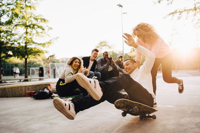 Laughing friends photographing man falling from skateboard while woman pushing him at park