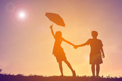 Full length of couple holding hands with umbrella on field against sky during sunset