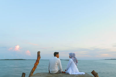 Rear view of couple sitting on pier over sea against sky during sunset