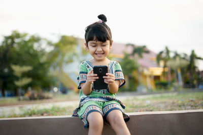 Girl using phone while sitting on retaining wall against sky