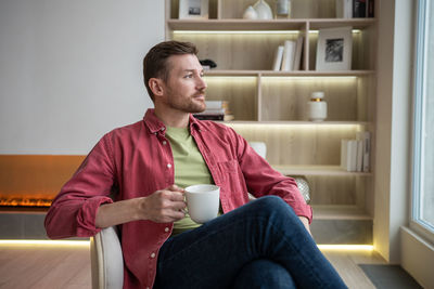 Calm positive man sitting in modern apartment with cup of tea, thinking, resting, looking at window