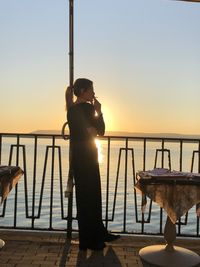 Woman smoking cigarette while standing by railing against sea and sky during sunset