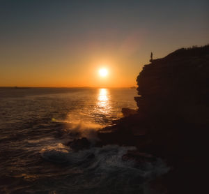 Drone aerial of silhouette of person standing at coastal cliff edge during golden hour sunset