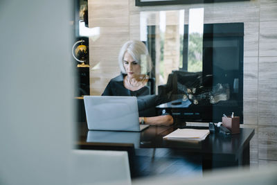 Serious businesswoman working on laptop computer in office seen through door