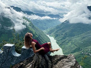 Woman sitting on rock looking at mountains against sky