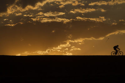 Silhouette person cycling against cloudy sky during sunset