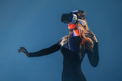 Side view of young unrecognizable blonde woman standing touching air and wearing virtual reality headset on colorful blue studio background