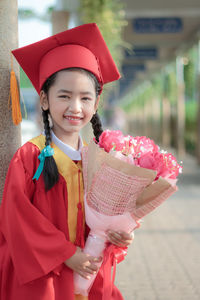 Portrait of smiling girl in graduation gown holding flowers while standing outdoors