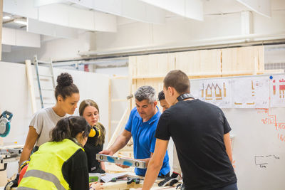 Male instructor using level amidst trainees at workbench in illuminated workshop