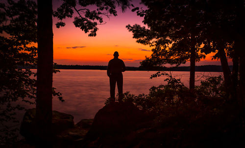 Silhouette man standing at lakeshore against orange sky