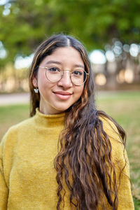 Content young female in eyeglasses looking at camera while sitting on grassy lawn in park with trees on blurred background