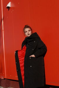 Fashionable teenage girl wearing black warm clothing standing against red wall during sunny day