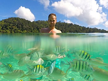 Young man standing amidst school of fish in sea against sky