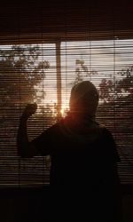 Teenage girl standing against window blinds during sunset