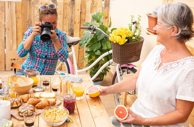 Woman photographing breakfast on table with mother holding oranges