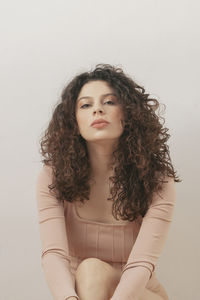 Gentle dreamy female with curly hair and in bodysuit sitting on floor against white wall in studio