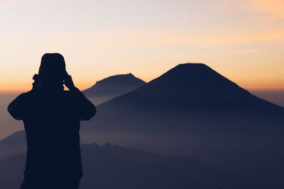 Silhouette man photographing mountains against sky during sunset
