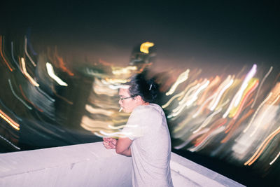 Side view of man smoking while standing on terrace against illuminated city at night