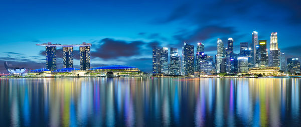 Illuminated modern buildings by sea against sky at night