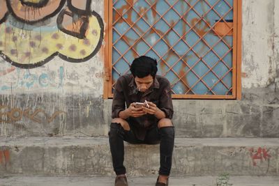 Full length of young man using phone while sitting on wall