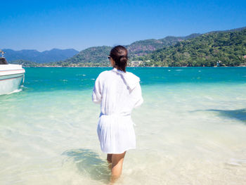 Rear view of woman standing on sea shore against mountains