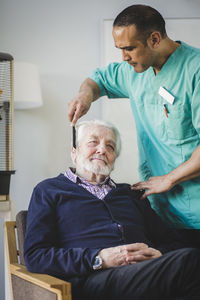 Mature male caregiver combing hair of senior man sitting on chair in nursing home