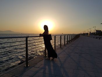Woman standing on railing against sea during sunset