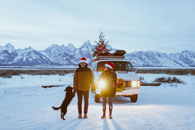 Couple laugh and hold hands in the tetons with dog and festive van christmas tree