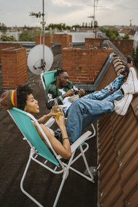 Full length of female and male friends sitting with feet up on chair at rooftop