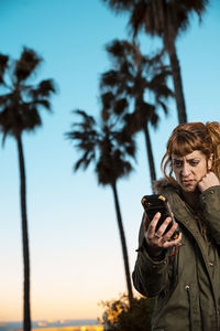 Woman using phone while standing against palm trees during sunset