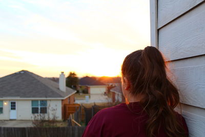 Rear view of woman leaning on wooden wall against clear sky during sunset