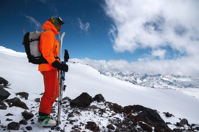 A young tall professional skier holds his skis while standing high in the mountains and looks 