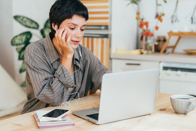 Young woman working on a laptop and listening to music on headphones at home in the kitchen.