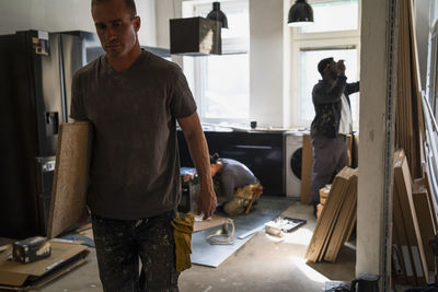 Male carpenter carrying plank while coworkers working in background in kitchen at apartment