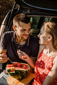 Millennial couple sitting on open trunk and eating watermelon. happy young couple having break at