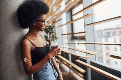 African girl with curly black hair and in casual clothes standing indoors with cup of drink.