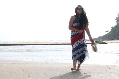 Full length of woman holding shoes while walking at beach against clear sky