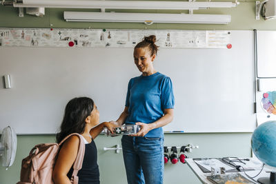 Smiling female teacher collecting smart phone of schoolgirl while standing in classroom at elementary school