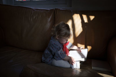 Girl reading book while sitting on sofa at home