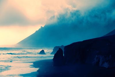 Scenic view of cliff by sea in foggy weather during sunset