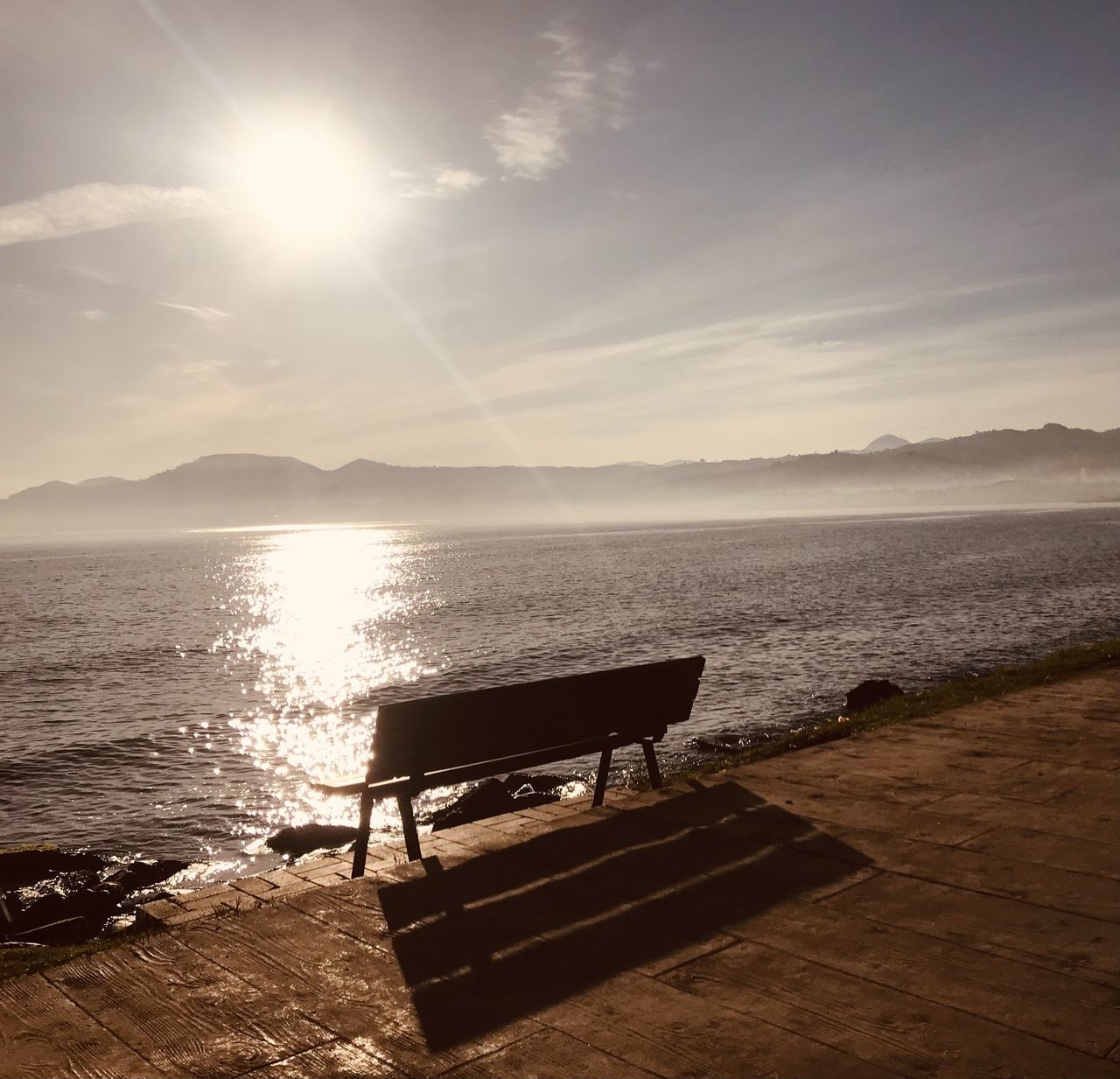 EMPTY BENCH ON SHORE BY SEA AGAINST SKY