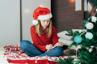 Full length of woman sitting by christmas tree at home