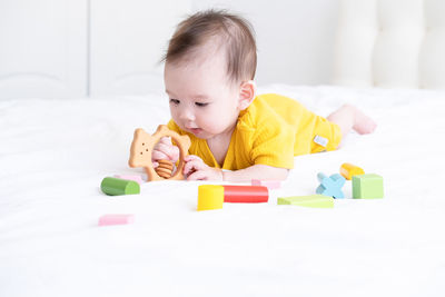 Healthy asian baby girl in yellow bodysuit playing with wooden toys on white bedding