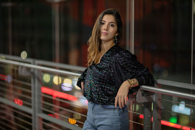 Portrait of young woman looking away while leaning against railing at night