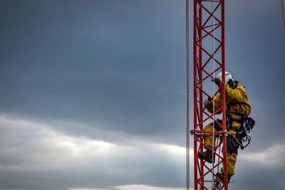 Low angle view of worker climbing built structure against cloudy sky